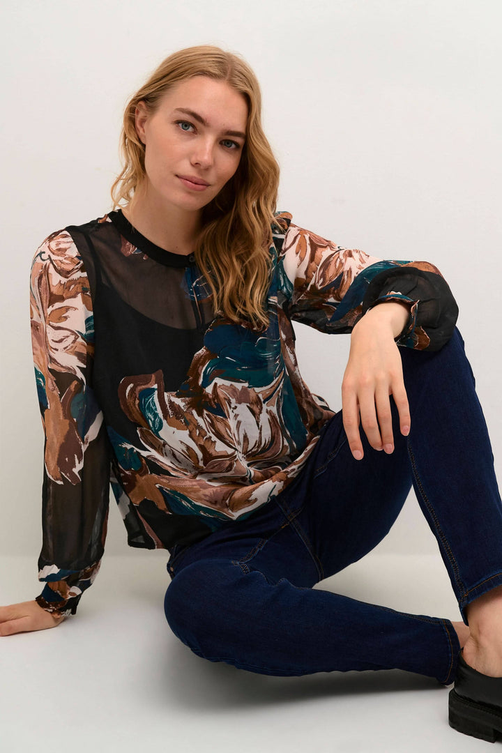 Femme portant la blouse Jasmina à imprimé floral avec manches longues, assise sur le sol, vêtue de jeans.