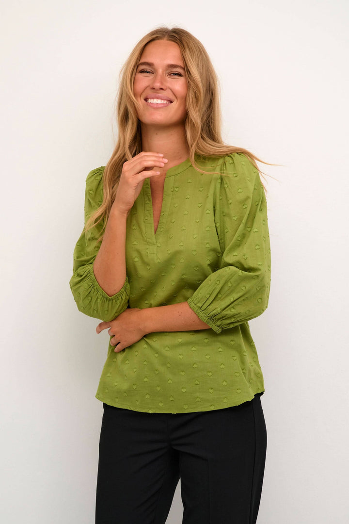 Femme souriante portant une blouse verte à manches 3/4 et encolure en V, parfaite pour un look décontracté.