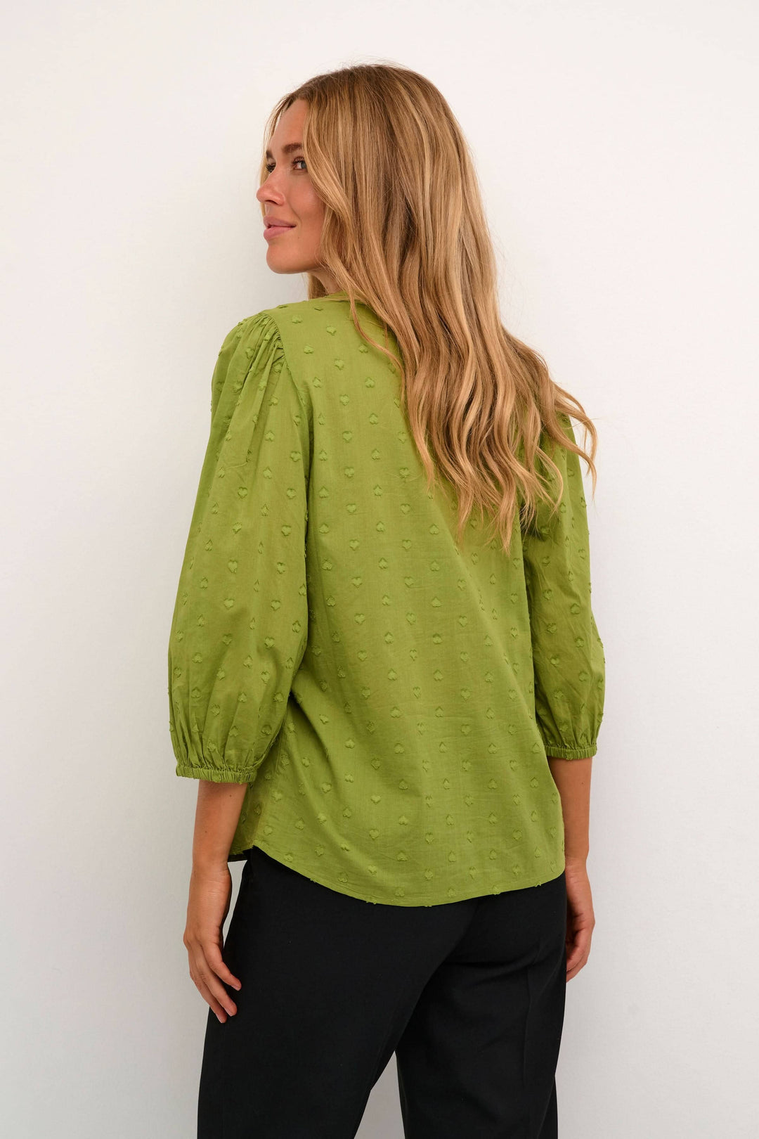 Femme portant une blouse verte à manches bouffantes, vue de dos, avec des détails texturés.