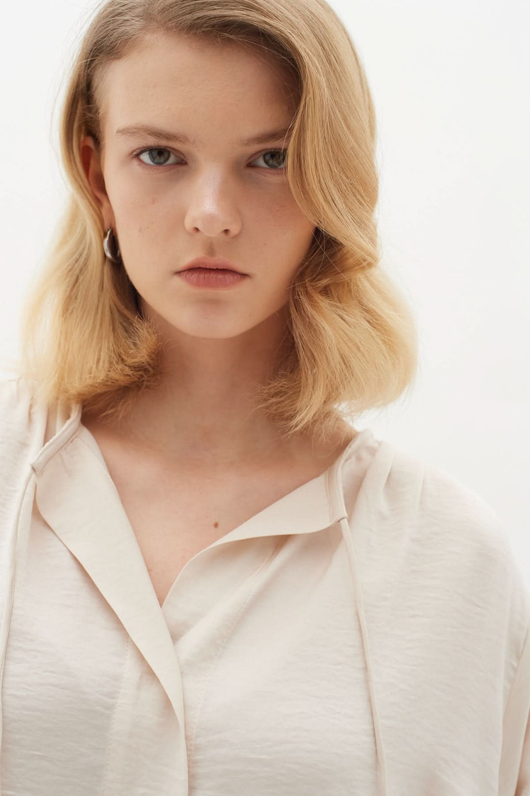 Femme avec un visage sérieux portant une blouse classique à encolure nouée de couleur crème.