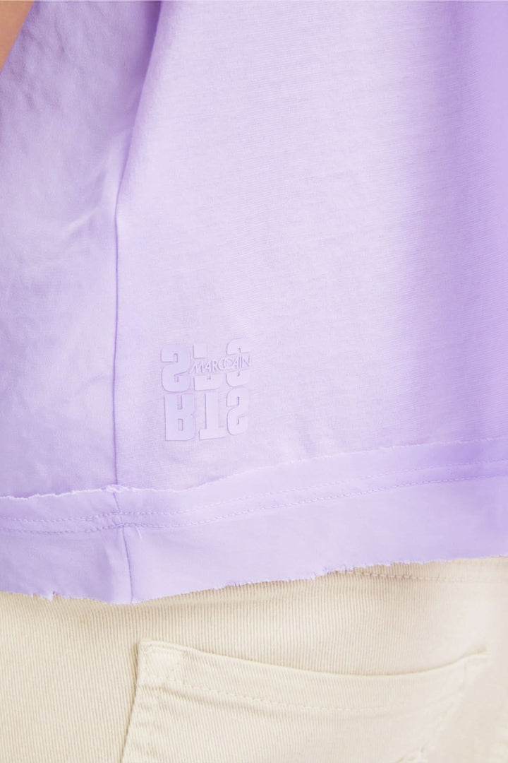 Détail d'une blouse lilas avec logo en relief et ourlet à bords bruts, portée sur un pantalon beige.