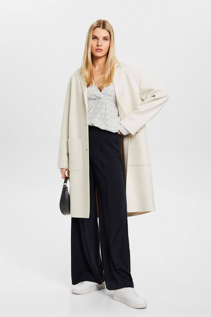 Femme en blouse col V et pantalon ample, portant un manteau beige, tenue décontractée et élégante.