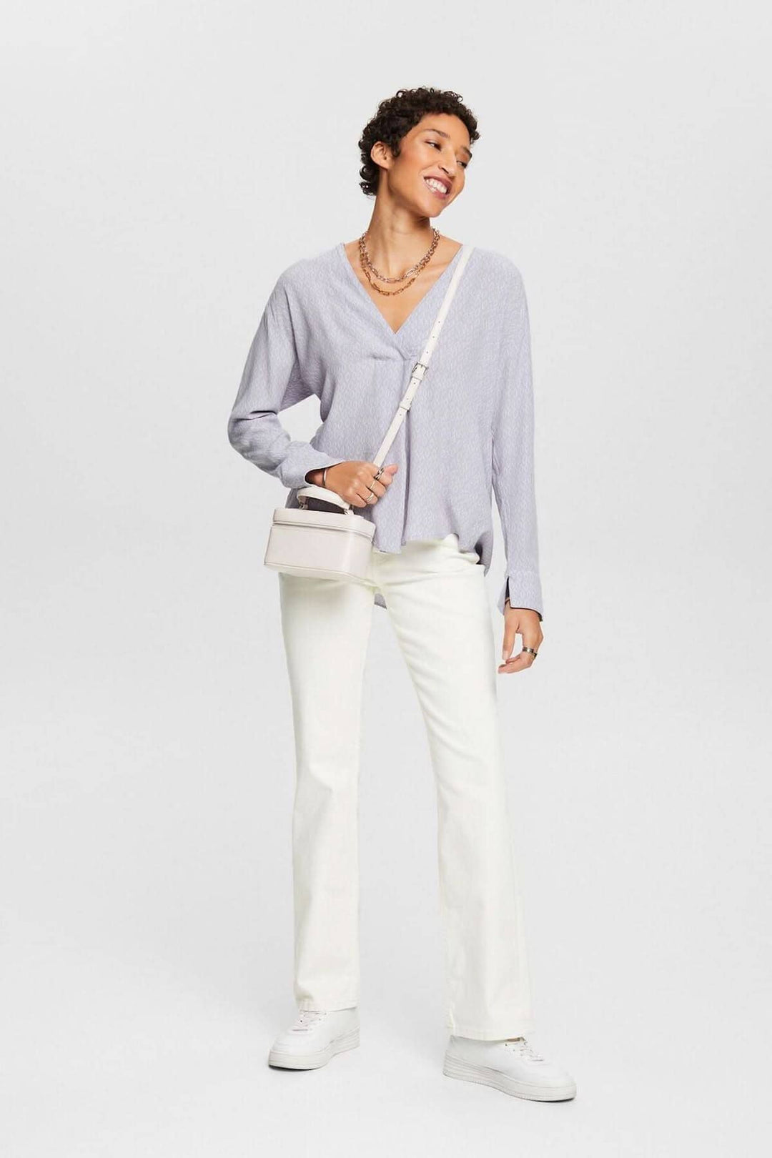 Femme souriante portant un chemisier col V en crêpe texturé avec pantalon blanc et sac à main.
