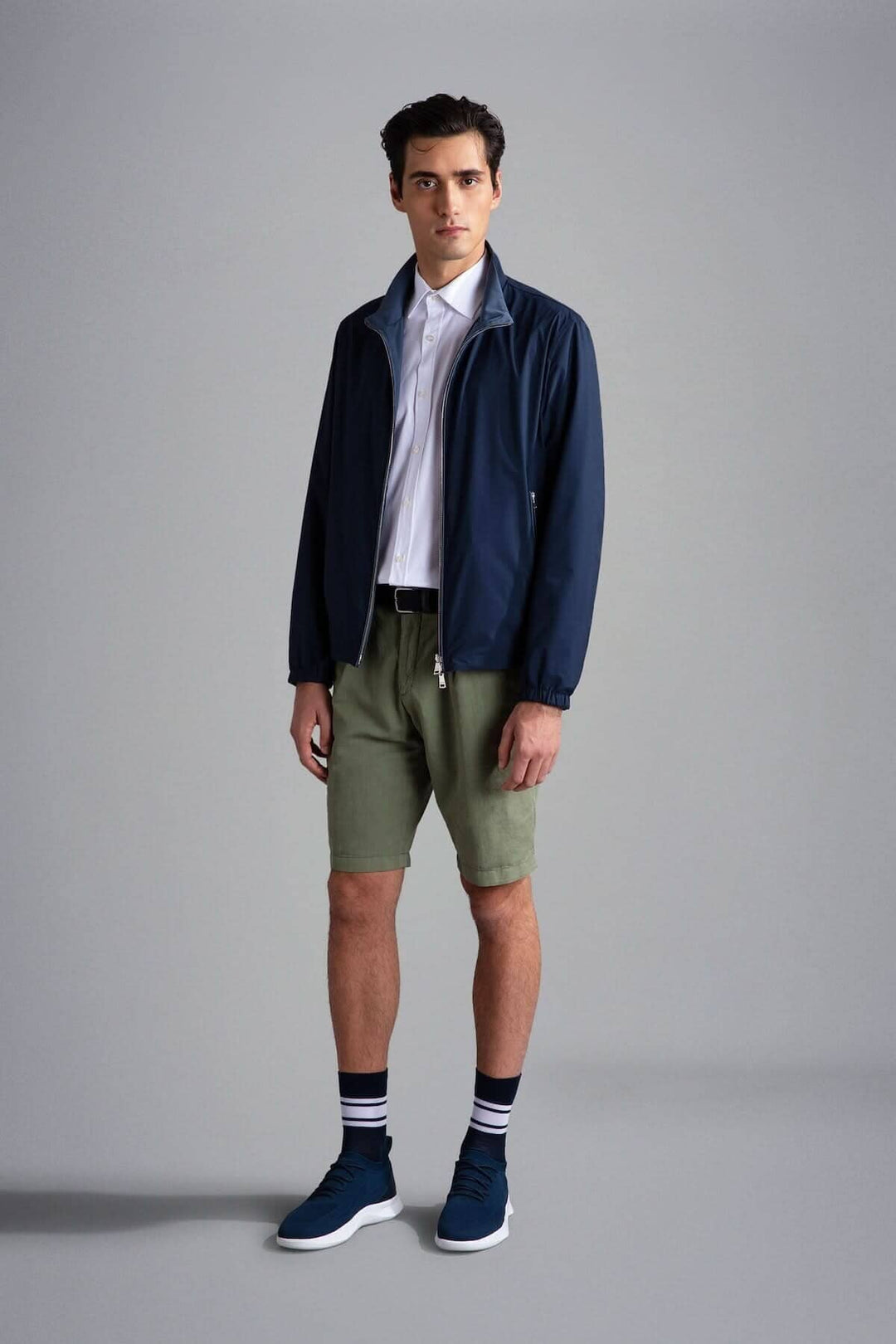 Homme en blouson imperméable réversible, chemise blanche, shorts olive et baskets bleues.