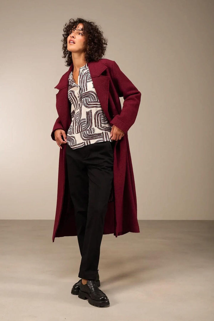 Femme portant une blouse à manches 3/4 avec encolure ronde et pantalon, style moderne et élégant dans un manteau bordeaux.