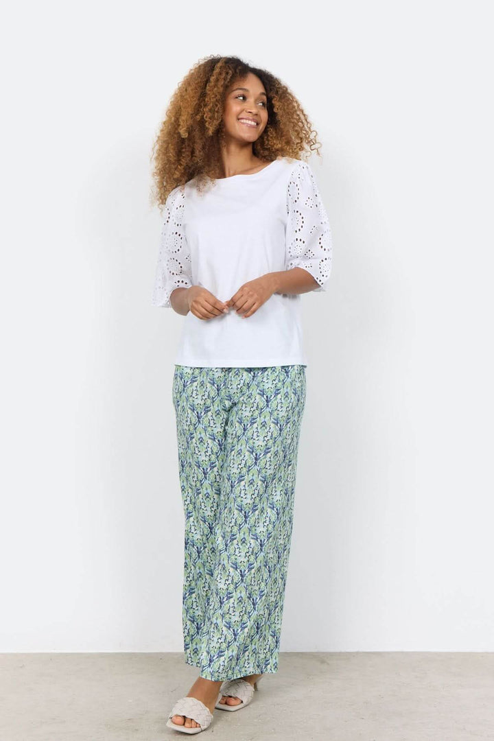 Femme souriante portant une blouse blanche à manches brodées et un pantalon long à motifs colorés.