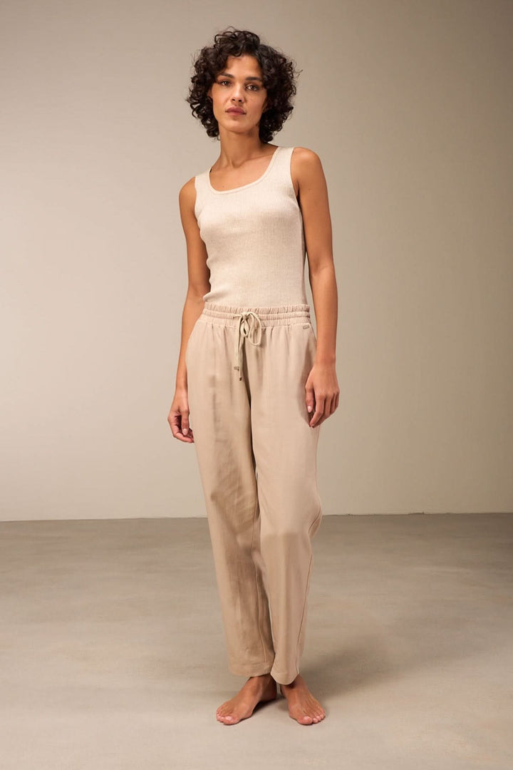 Femme en camisole beige sans manches et pantalon léger, posant sur un fond neutre.