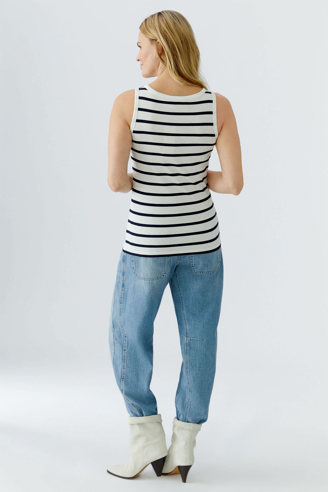 Femme portant une camisole rayée sans manches au style marin revisité, vue de dos, associe des chaussures blanches et un jean.