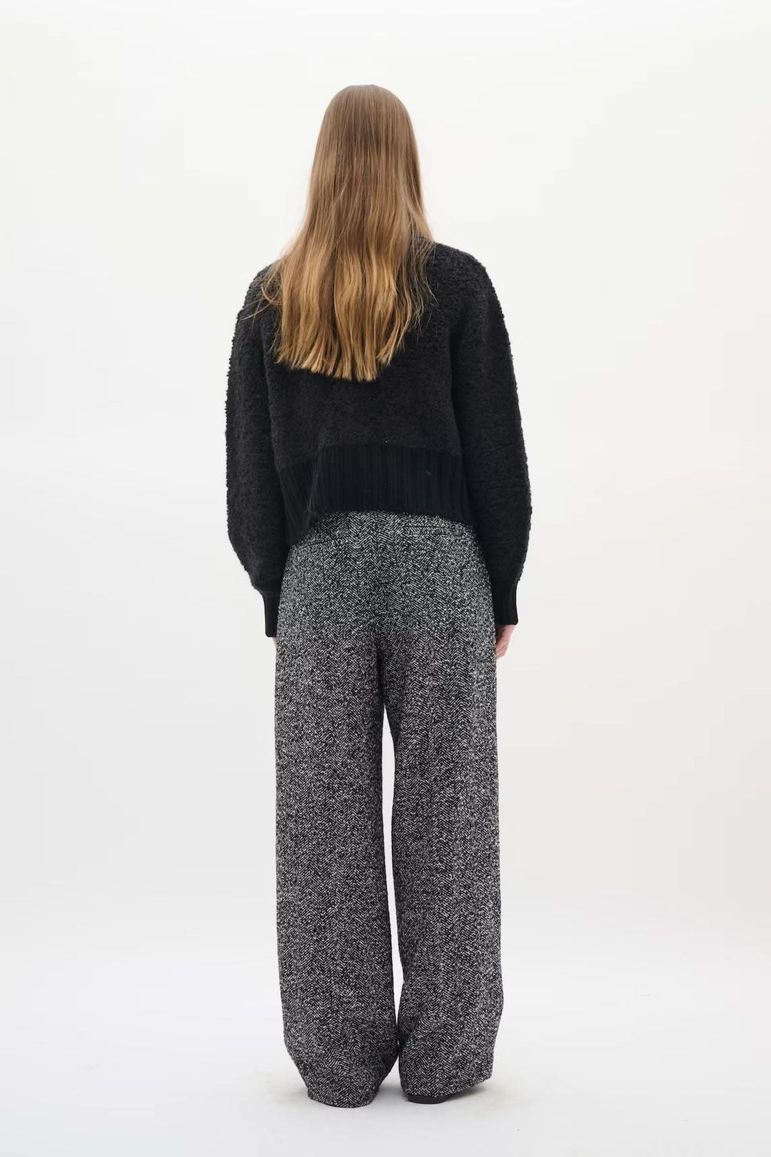 Person wearing Cardigan Guo, black textured knit with golden buttons, styled with high-waist pants for a chic silhouette from the back.