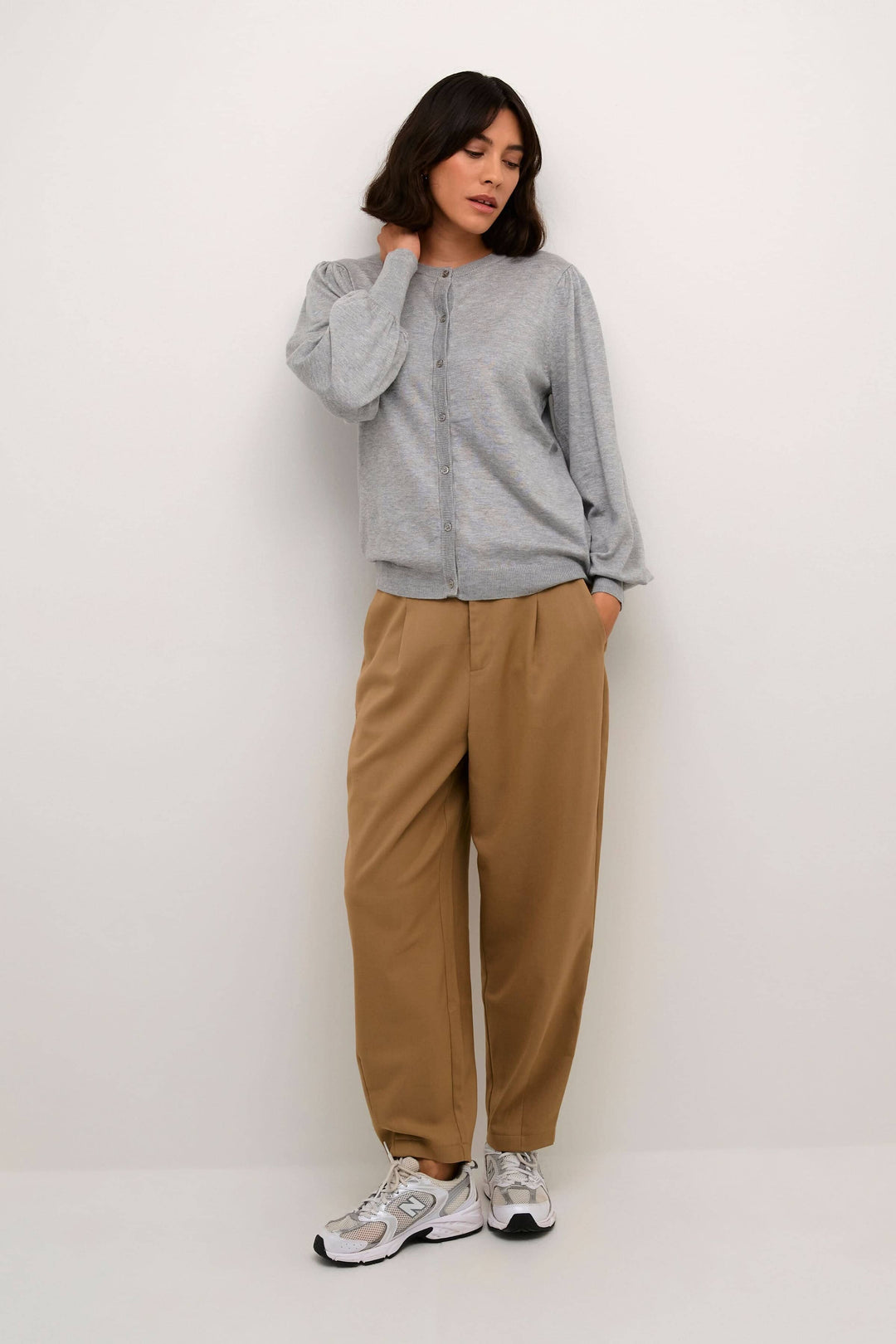 Cardigan Lizza gris à manches longues, col rond, fermé par des boutons, porté avec un pantalon beige et des sneakers.