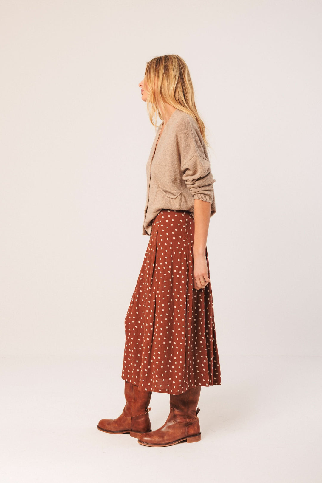 Femme portant un cardigan court beige, une jupe longue à pois et des bottes marron, style décontracté chic.