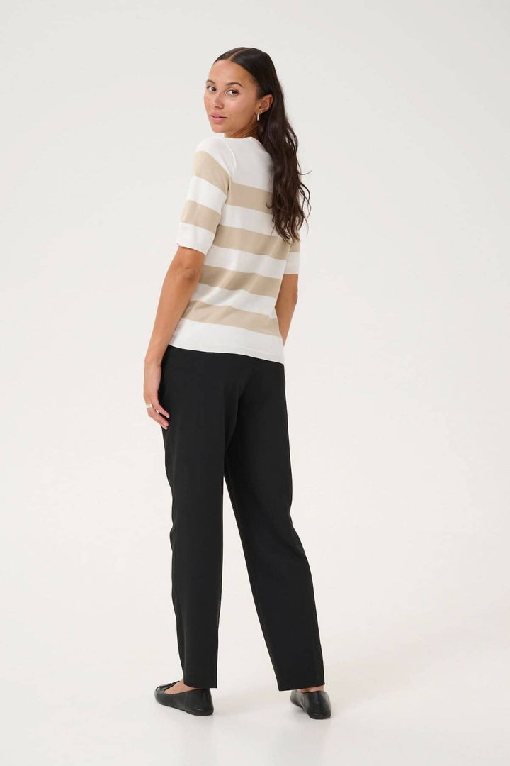 Femme portant un chandail ligné beige et blanc, dos tourné, avec pantalon noir, dans un style décontracté élégant.