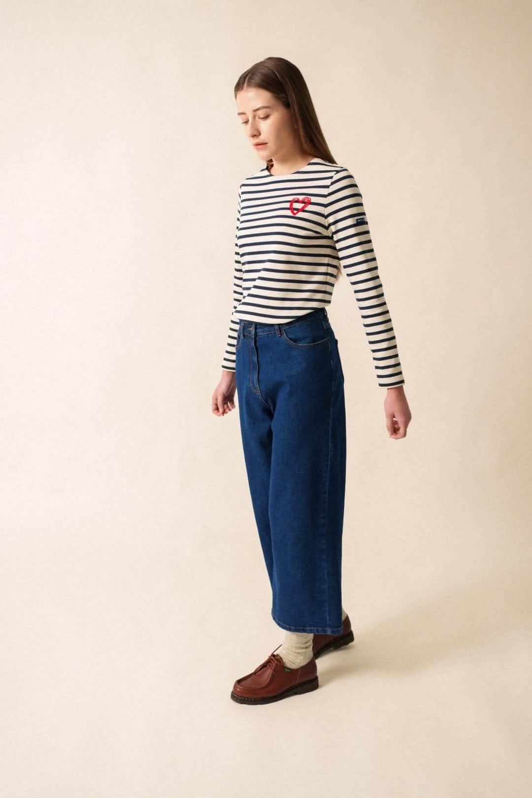 Femme en chandail rayé avec motif cœur, portant un jean bleu pour un look casual-chic.