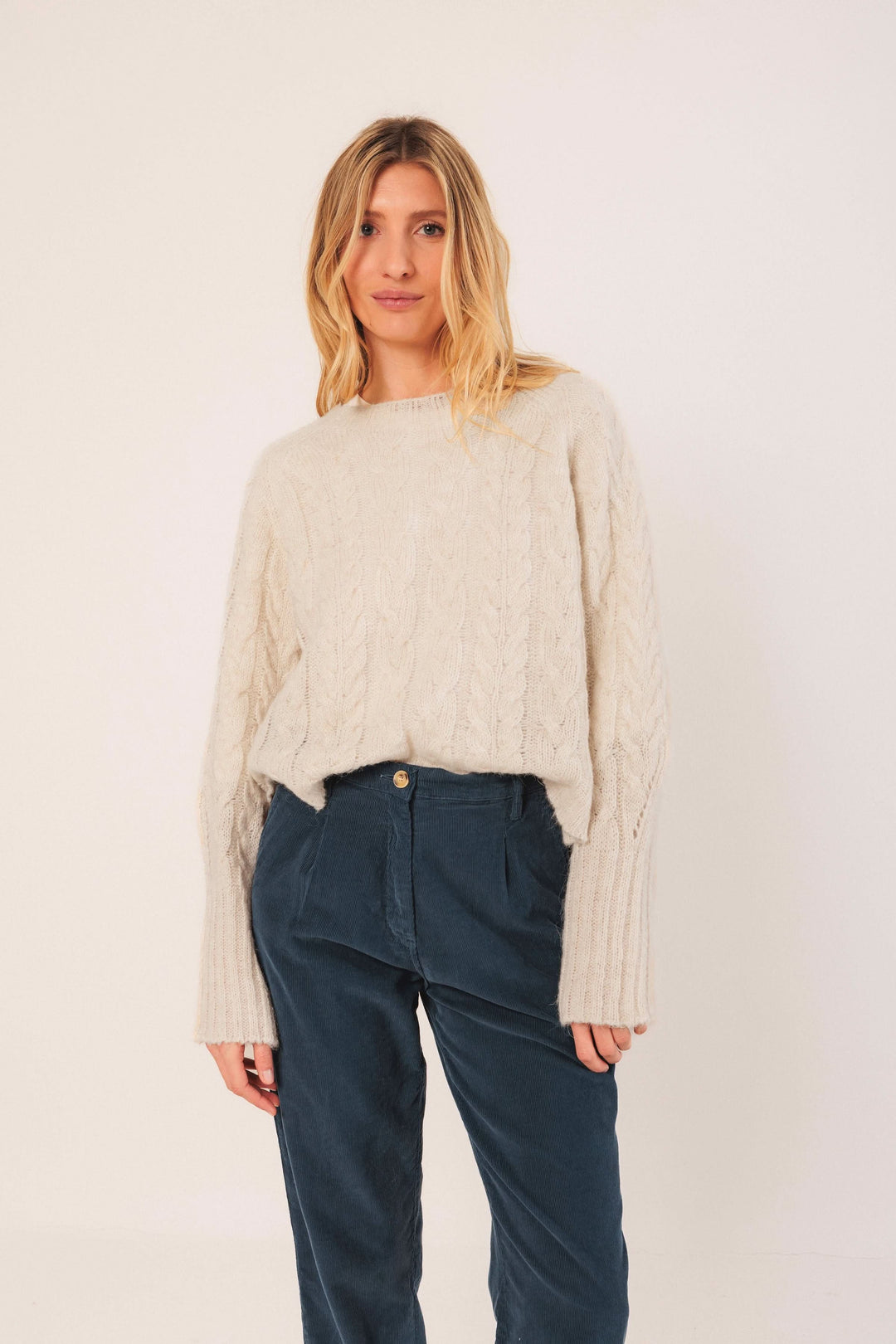 Chandail en maille torsadée beige, col rond, ample, porté avec pantalon bleu pour un look décontracté et élégant.
