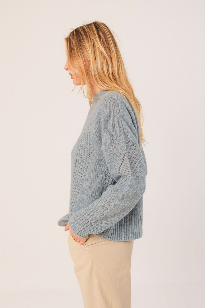 Chandail en maille légère bleu avec col rond, coupe droite et détails ajourés, porté avec un pantalon beige.