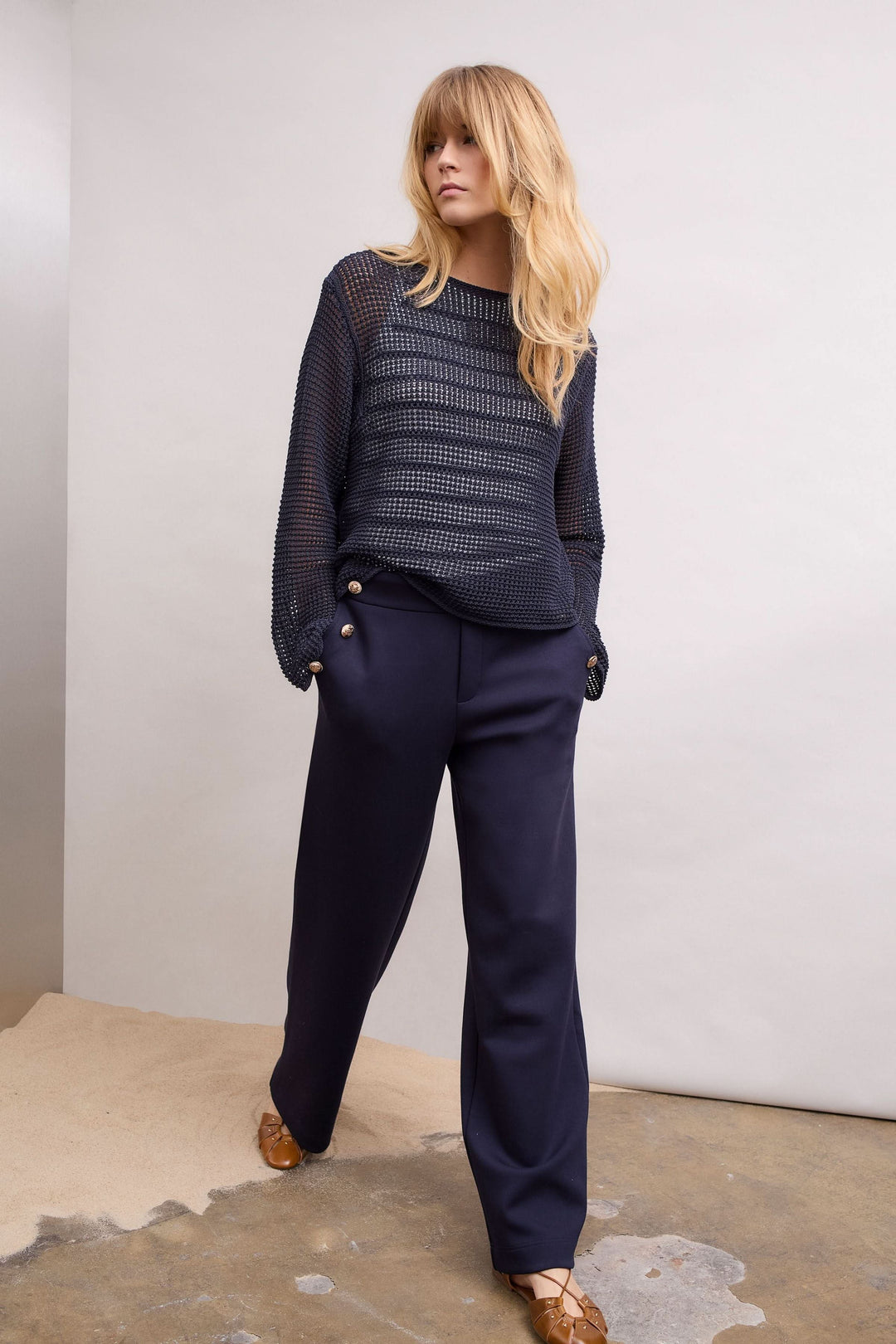 Femme portant un chandail en maille ajourée à manches longues et un pantalon bleu, pose élégante et moderne.