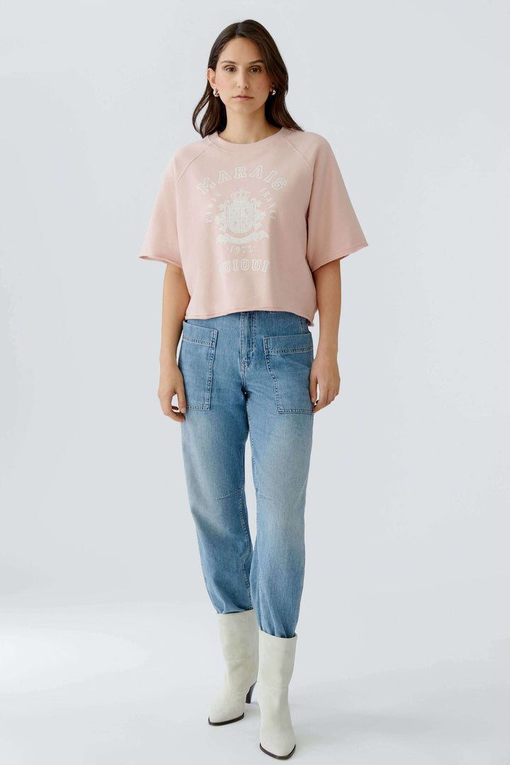 Femme portant un chandail imprimé graphique rétro avec coupe ample et manches courtes, combiné à un jean décontracté et des bottes blanches.