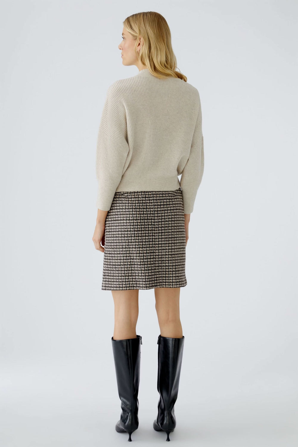 Femme de dos portant un chandail en maille fine et une jupe à motifs, avec des bottes noires élégantes.