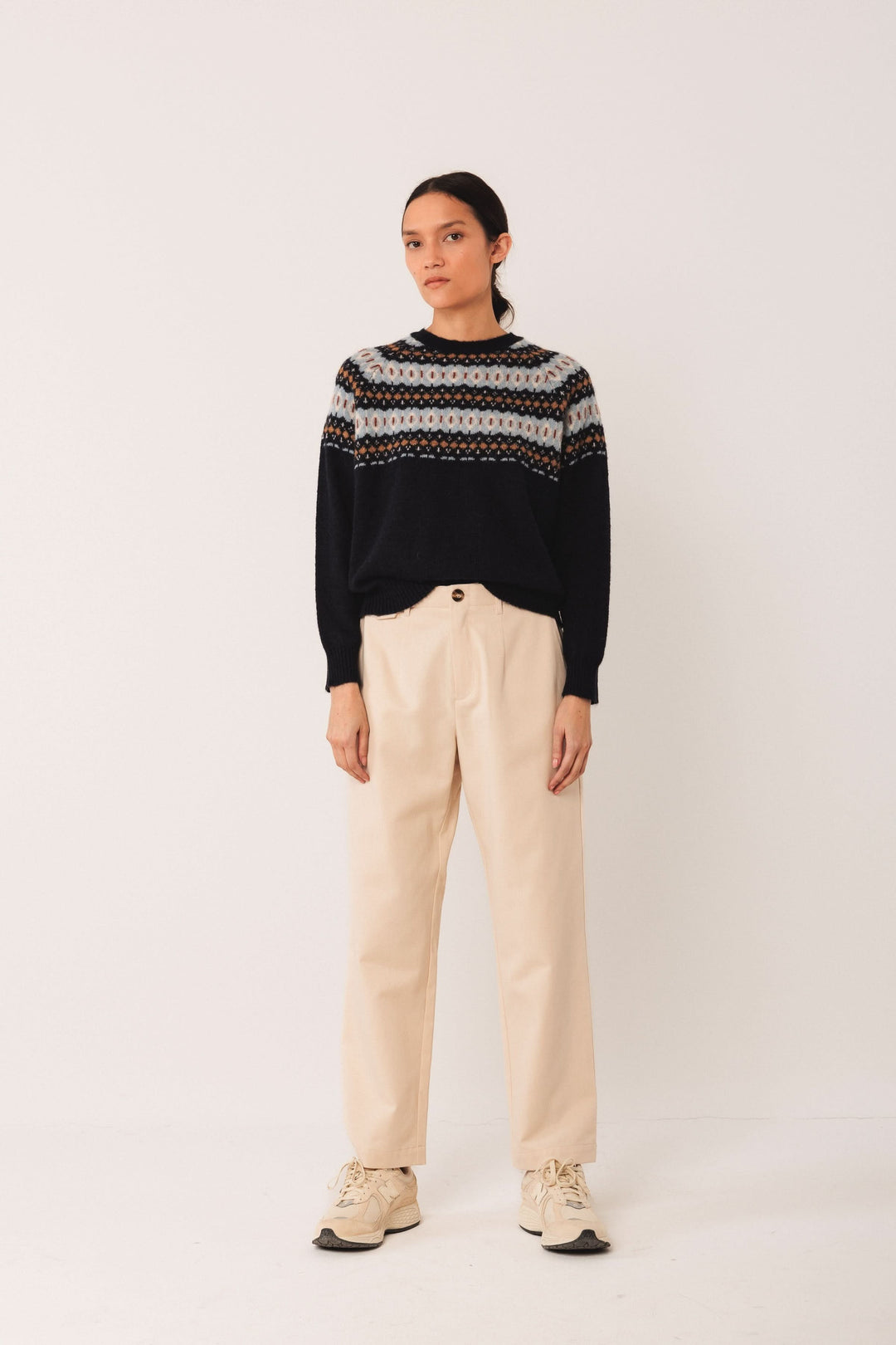 Femme portant un chandail jacquard noir avec motif coloré, associé à un pantalon beige, style décontracté.