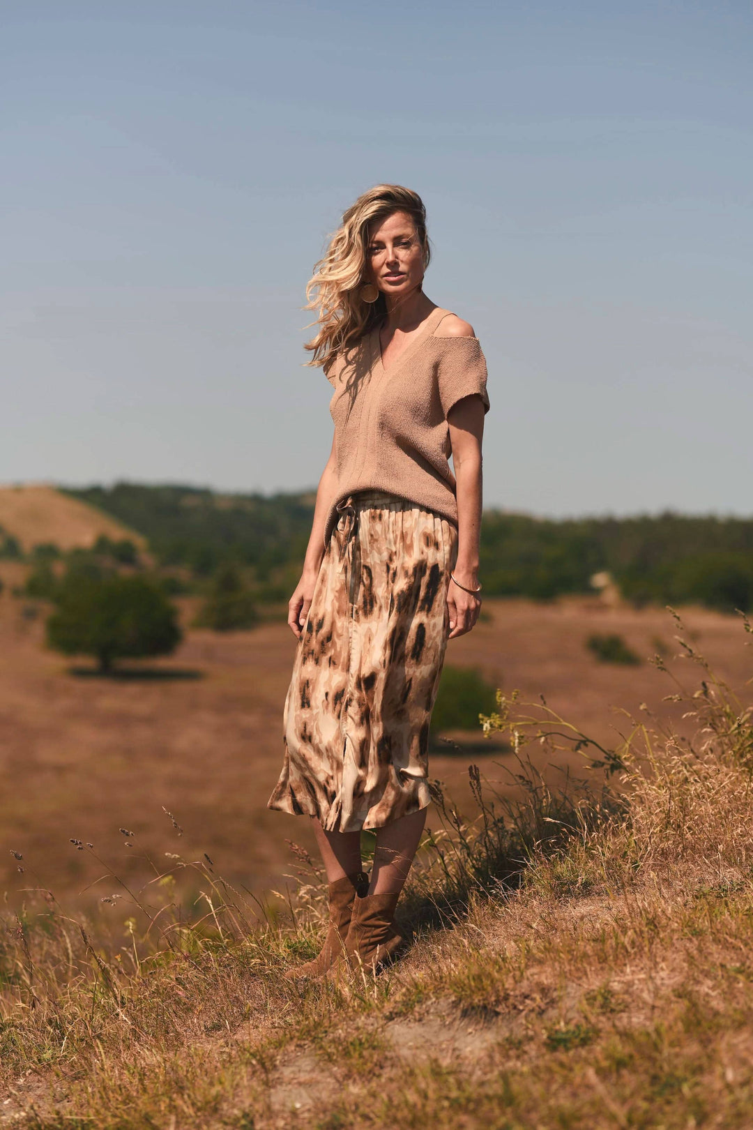 Femme portant un chandail sans épaules beige et une jupe imprimée, posant en extérieur sur un terrain naturel.