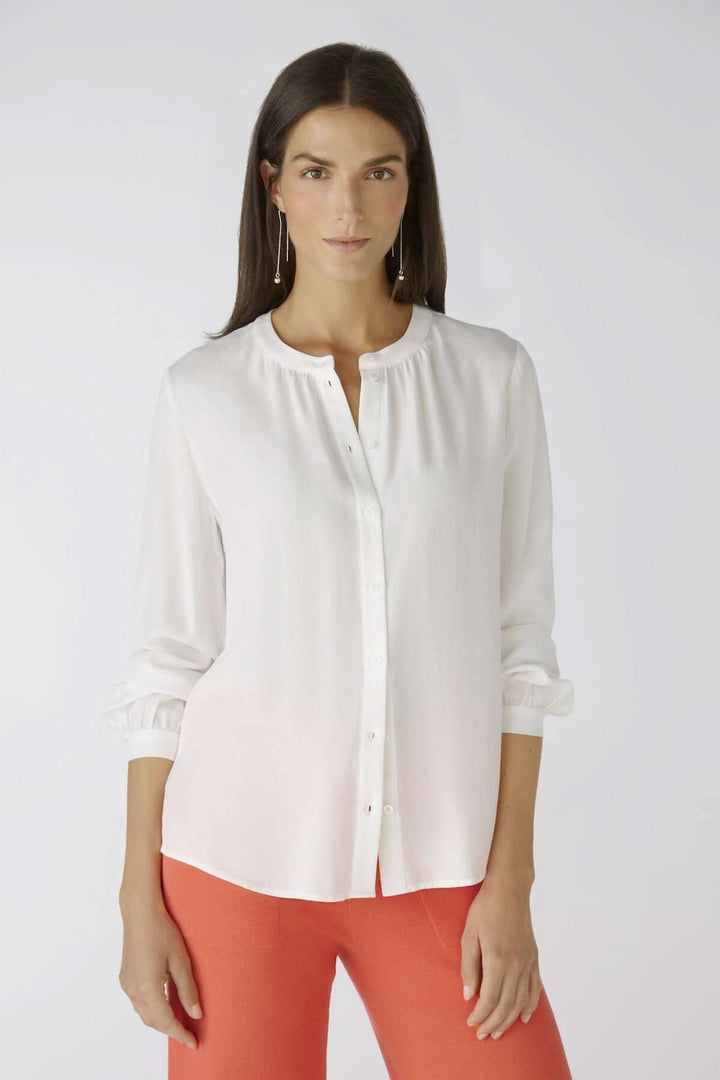 Chemise sans col blanche avec détails raffinés, manches longues, et encolure ronde, portée avec un bas coloré.
