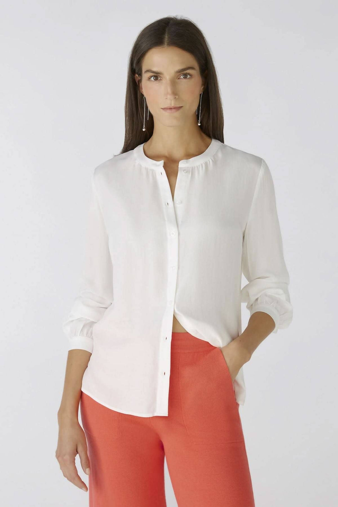 Chemise blanche sans col avec détails de boutons et manches longues, portée avec un pantalon rouge.