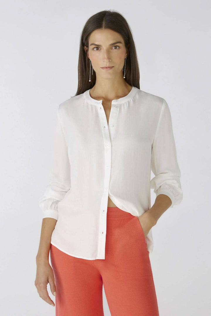 Chemise blanche sans col avec détails de boutons et manches longues, portée avec un pantalon rouge.