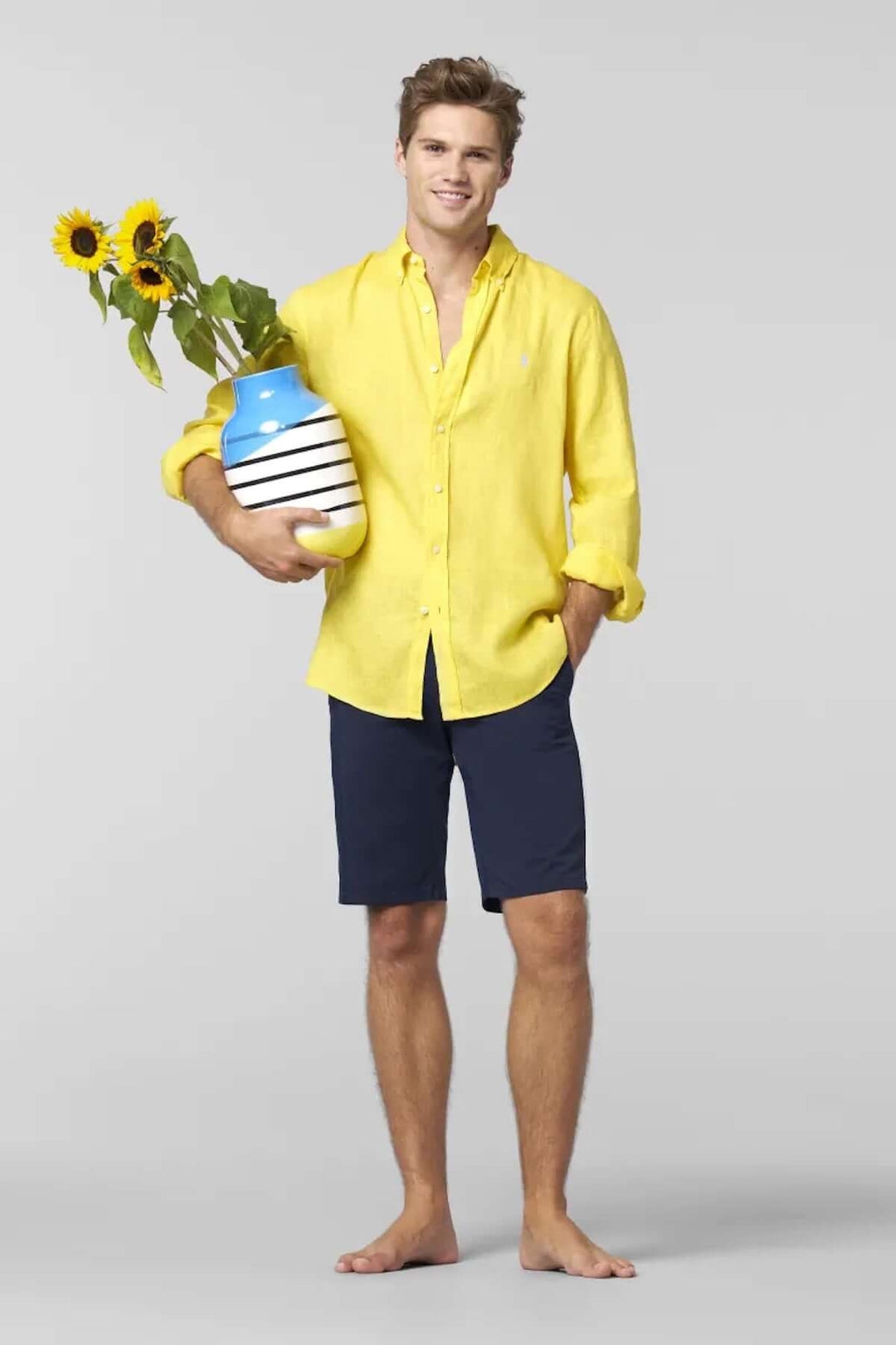Homme portant un chemisier jaune et un short, tenant un vase avec des tournesols, style estival décontracté.