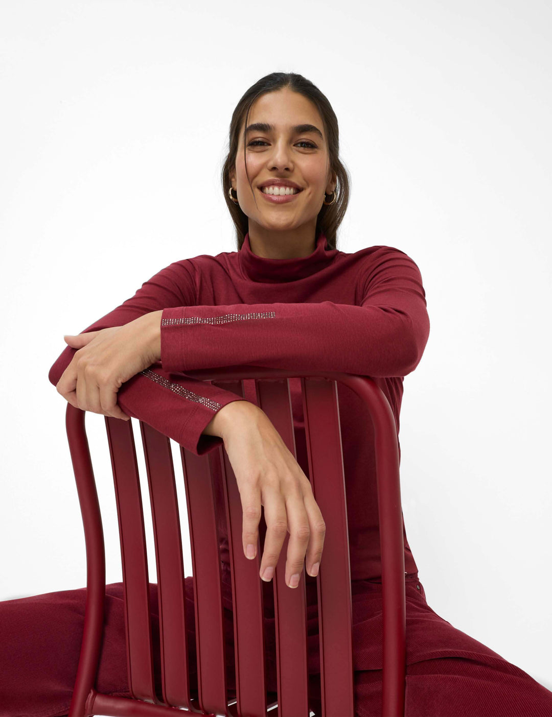 Femme portant un Col roulé CARINA.L bourgogne assise sur une chaise rouge, expression souriante, mode automne-hiver.