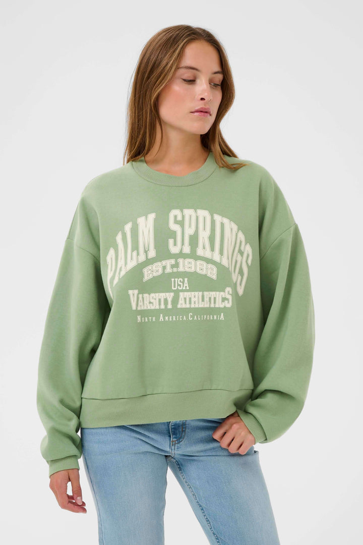 Jeune femme portant un coton ouaté Sussi vert avec motif universitaire, coupe ample et confortable, style décontracté et tendance.