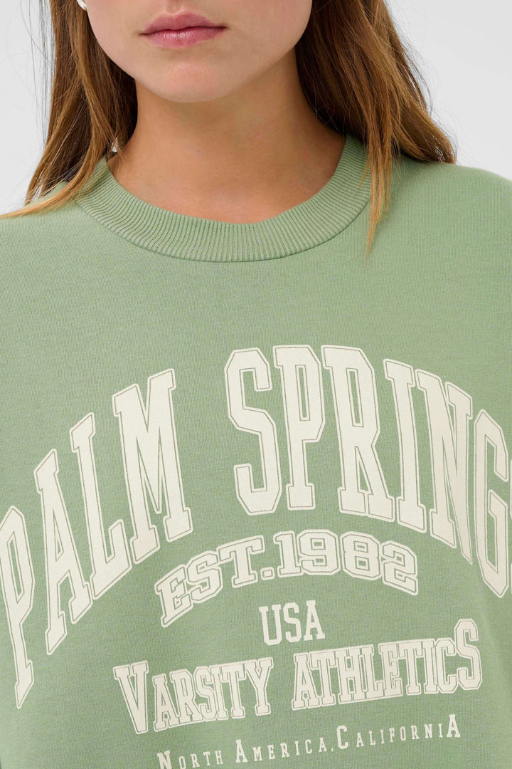 Coton ouaté Sussi vert à motif universitaire Palm Spring porté par un mannequin, offrant une coupe ample et confortable.