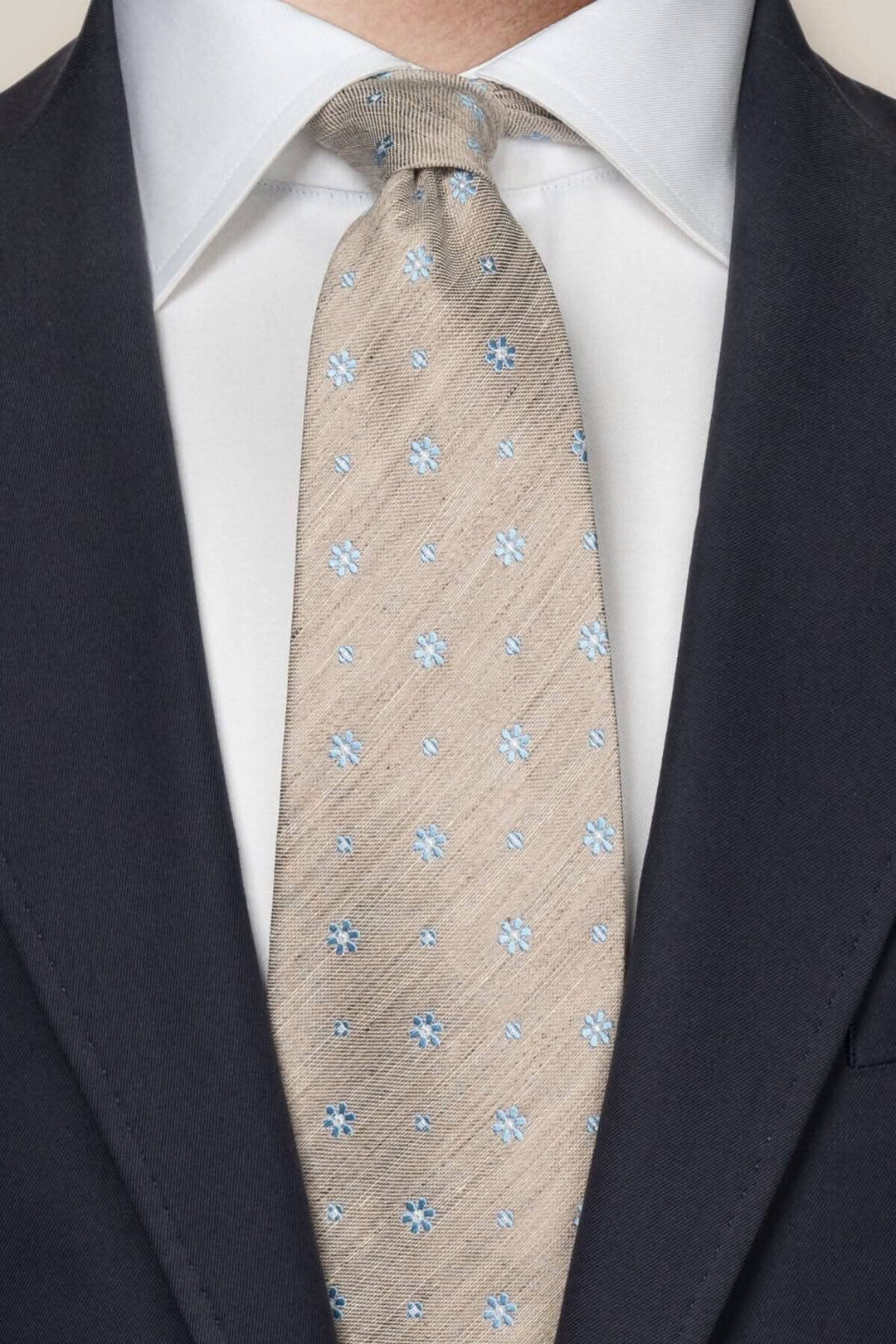 Cravate en soie et lin beige avec micro motifs floraux bleus, portée avec une chemise blanche et un costume noir.