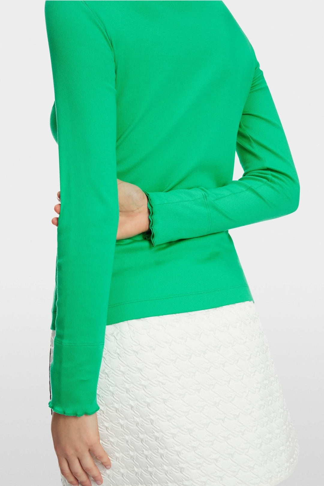Haut col mock ajusté vert avec manches longues et ourlets à bords bruts, porté avec une jupe blanche.