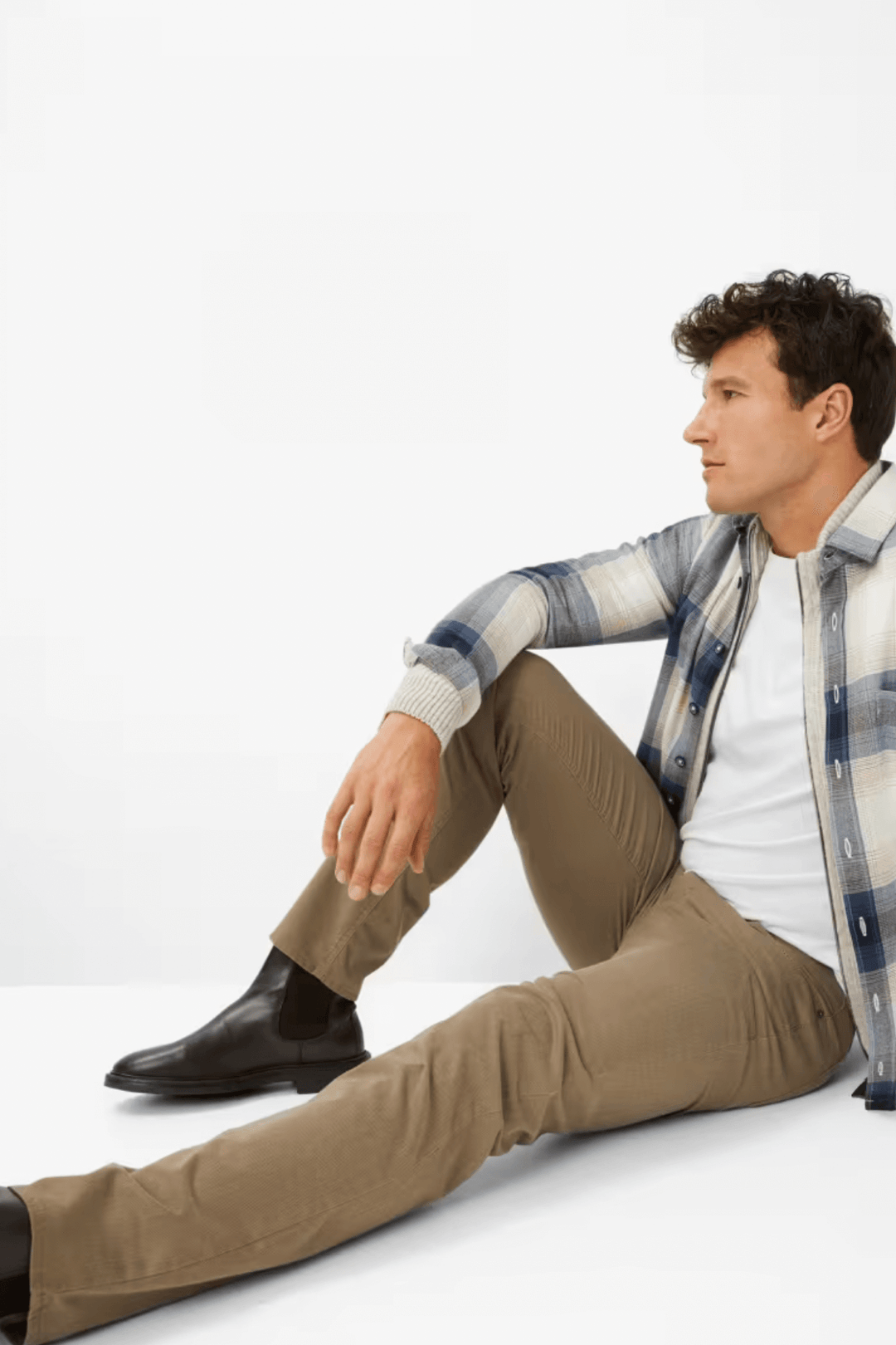 Homme en jean beige et chemise à carreaux, assis avec des bottes pour un look moderne et décontracté.
