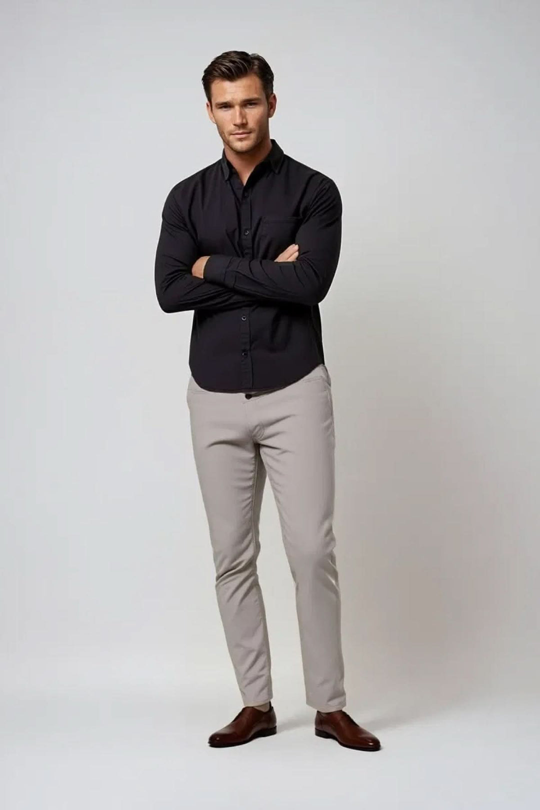 Homme en chemise noire et pantalons beiges, posant avec les bras croisés sur fond neutre.