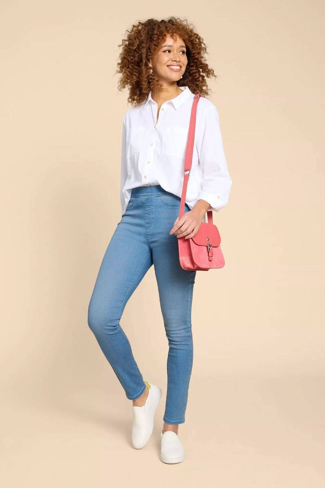 Femme souriante portant une chemise blanche, jeans ajustés et sac rose, pour un look décontracté élégant.