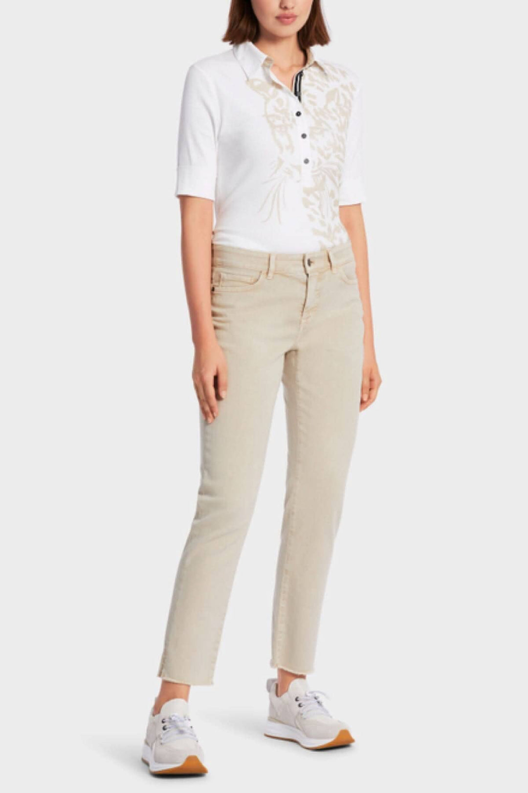 Femme portant un polo blanc à motifs et un jean beige, associé à des chaussures blanches, style décontracté.