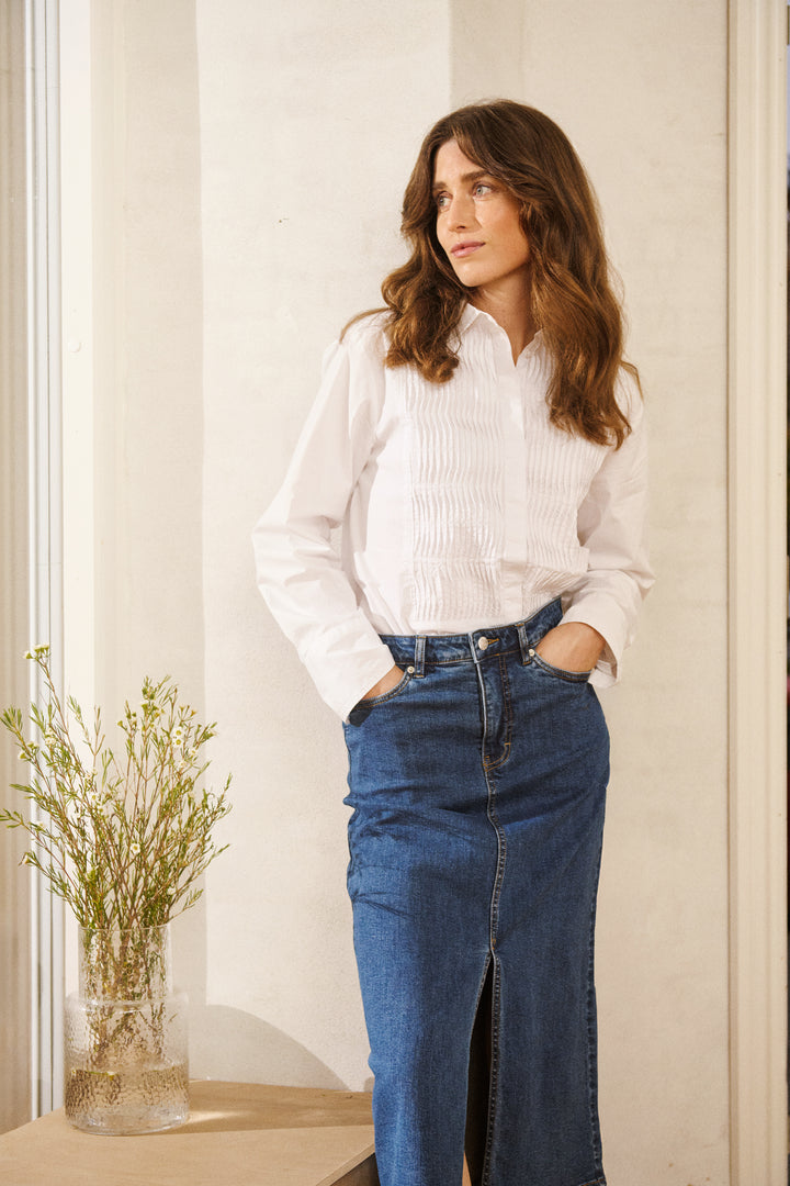 Femme debout en jupe en denim midi avec fente frontale et chemise blanche, look chic et décontracté.