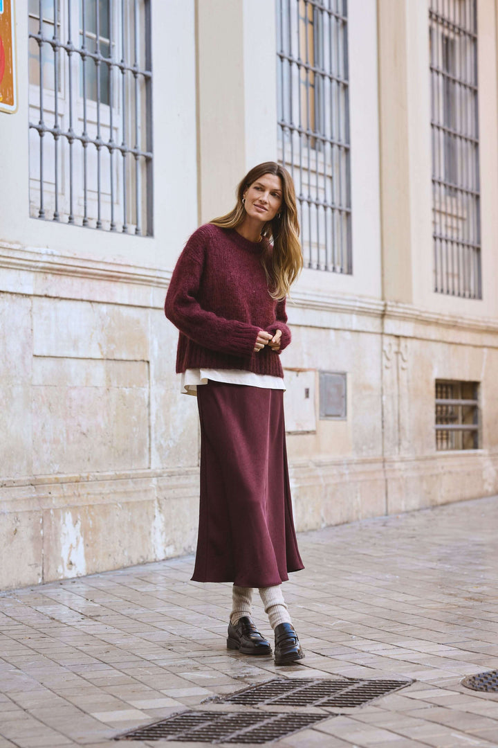 Femme portant une jupe midi fluide bordeaux et un pull oversize dans une rue élégante.