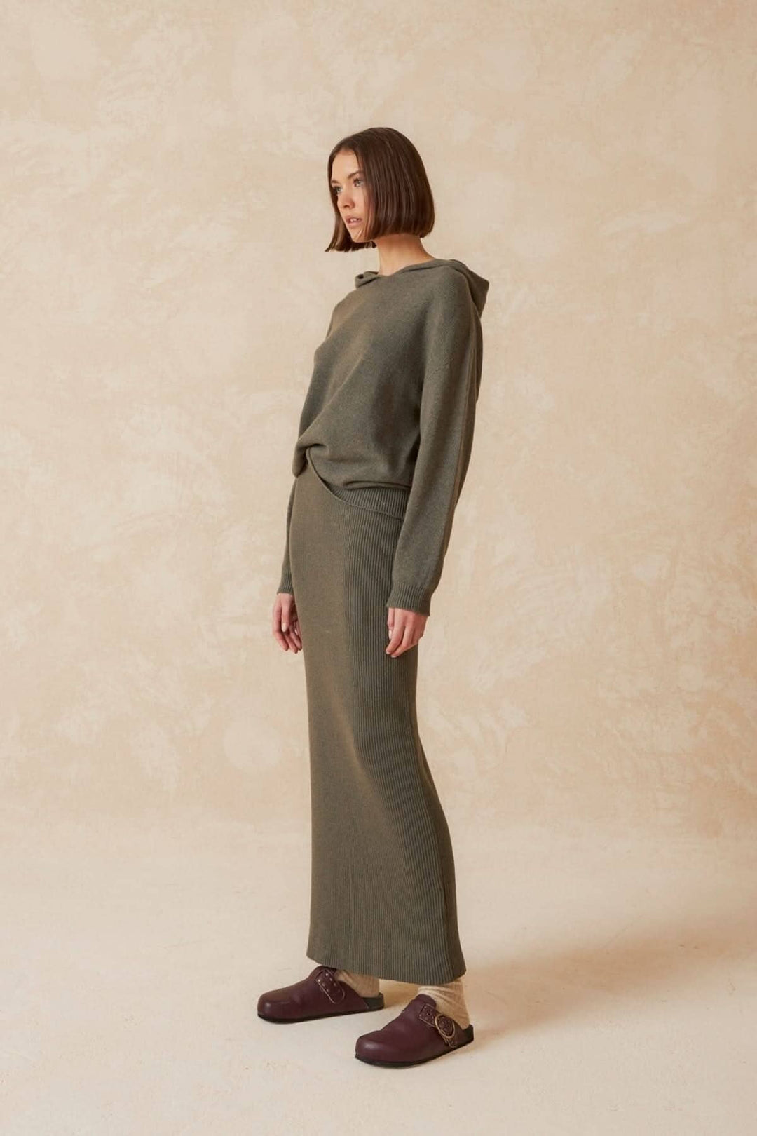 Femme portant une jupe longue verte avec un haut décontracté, associée à des clogs, sur fond minimaliste.