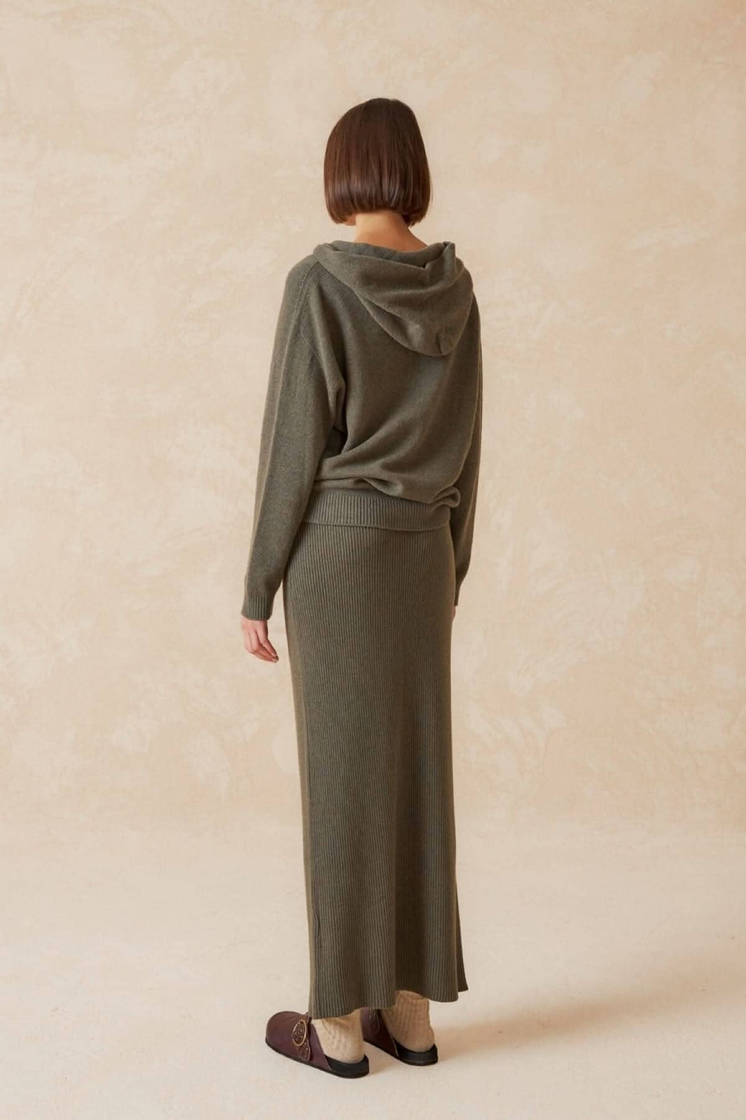 Femme de dos portant une jupe longue olive et un haut à capuche, style décontracté et élégant.