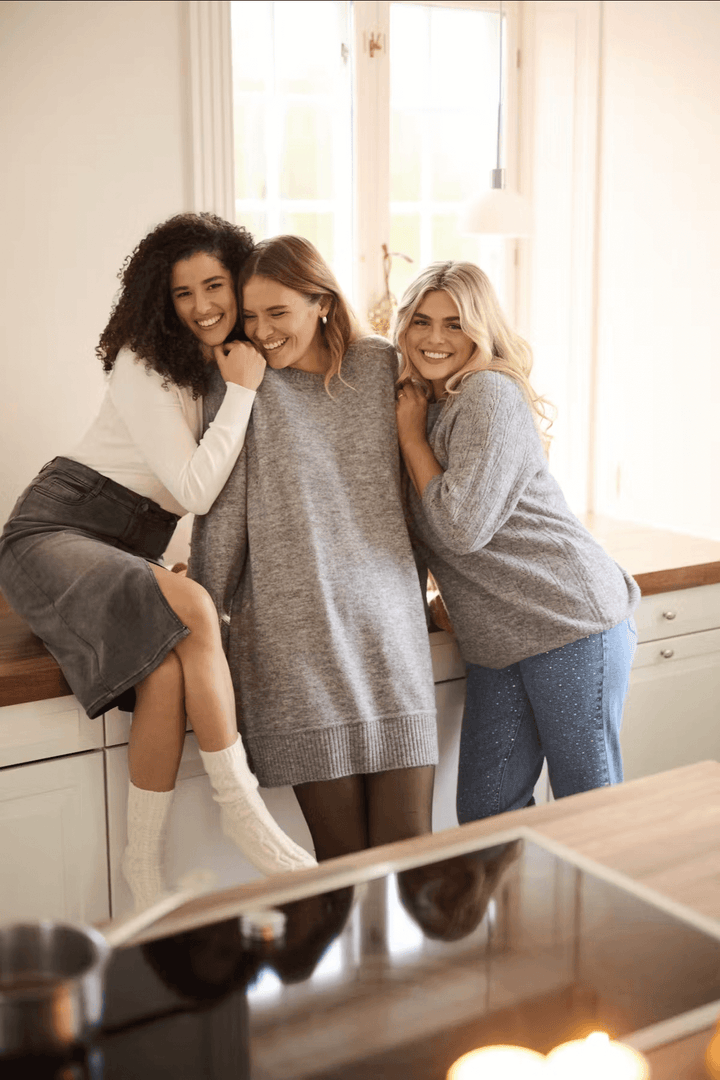 Trois femmes souriantes portant des pulls confortables, se tenant à un plan de travail dans une cuisine lumineuse.