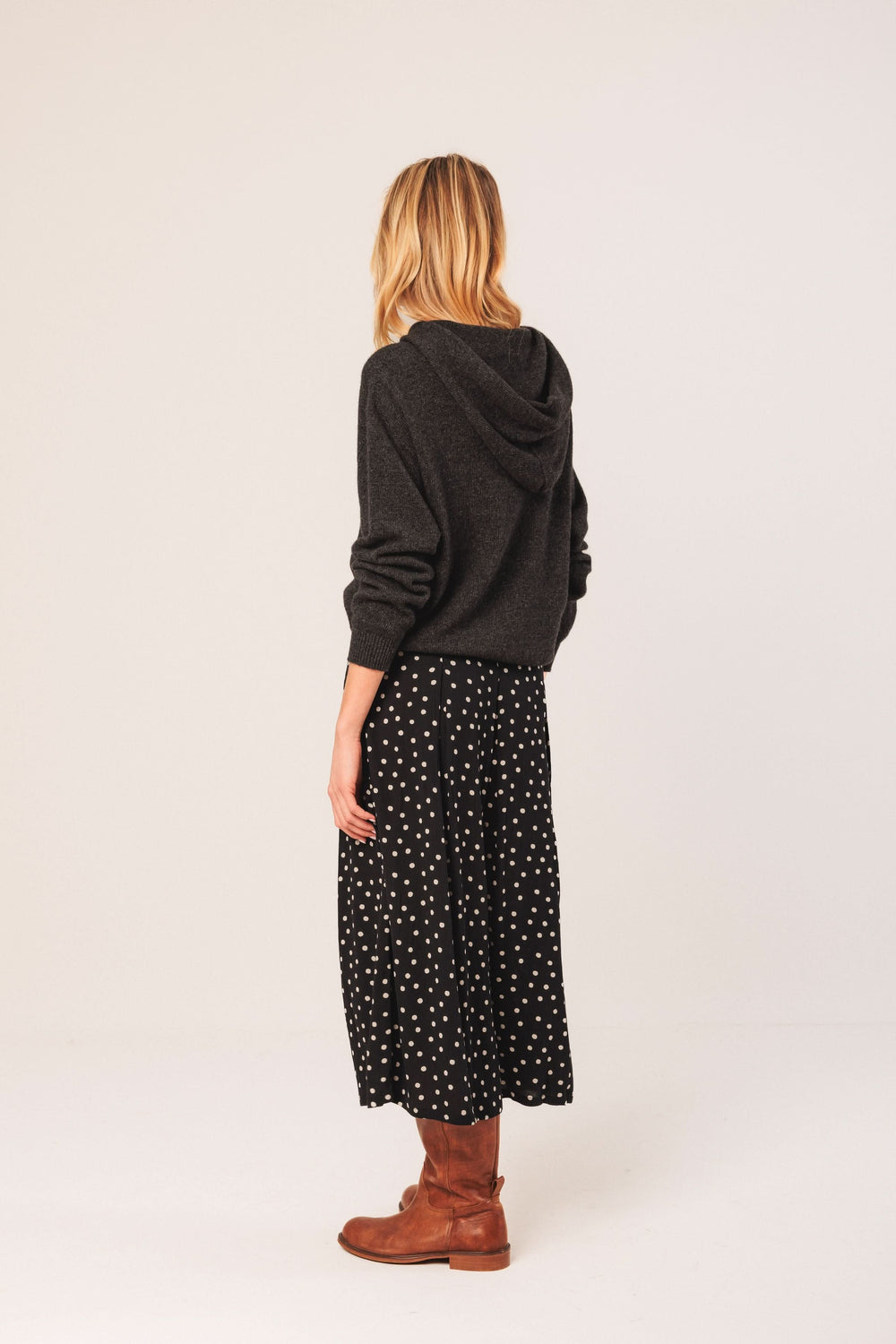 Femme de dos portant une jupe midi fluide à pois et un chandail gris, avec des bottes marron.