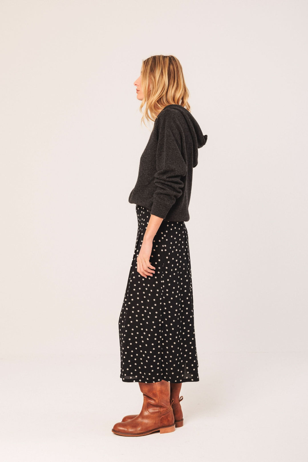 Femme en jupe midi à pois, associée à un chandail noir et des bottes marron, style décontracté.