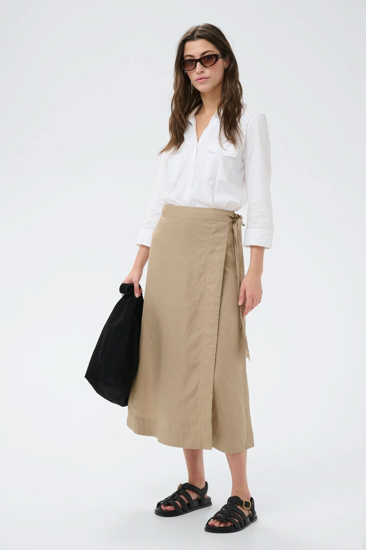 Femme portant une jupe porte-feuille Arbella beige avec chemise blanche, sac noir et sandales, silhouette fluide et élégante.