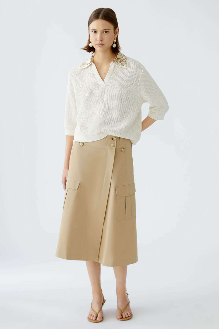 Modèle portant une jupe portefeuille beige en coupe trapèze avec un pull blanc et des sandales.