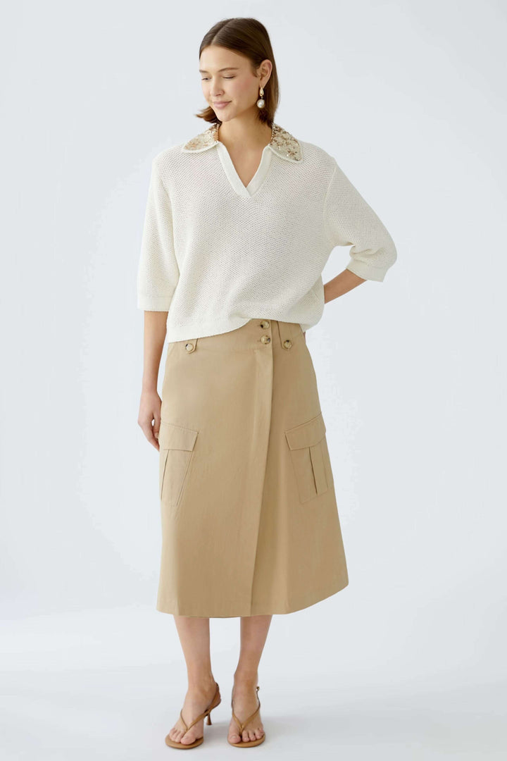 Femme portant une jupe portefeuille en coupe trapèze beige avec un pull blanc et des sandales, style moderne.
