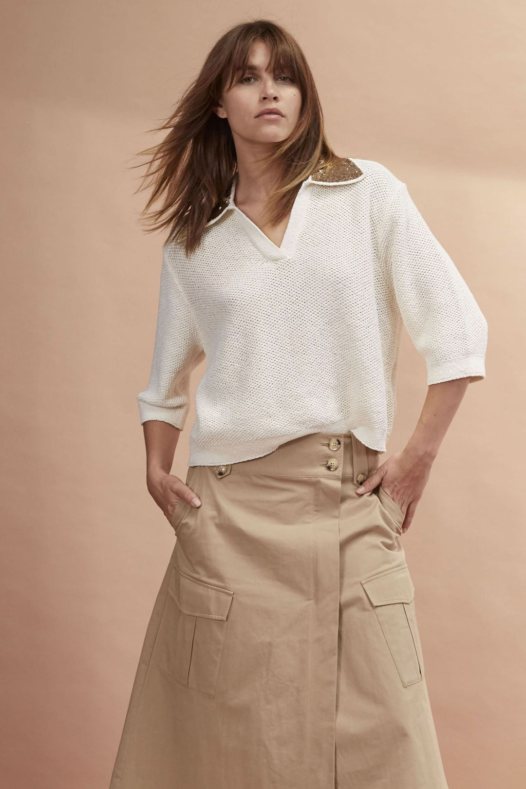 Femme en pull blanc avec col en cuir, portant une jupe portefeuille beige avec poches.