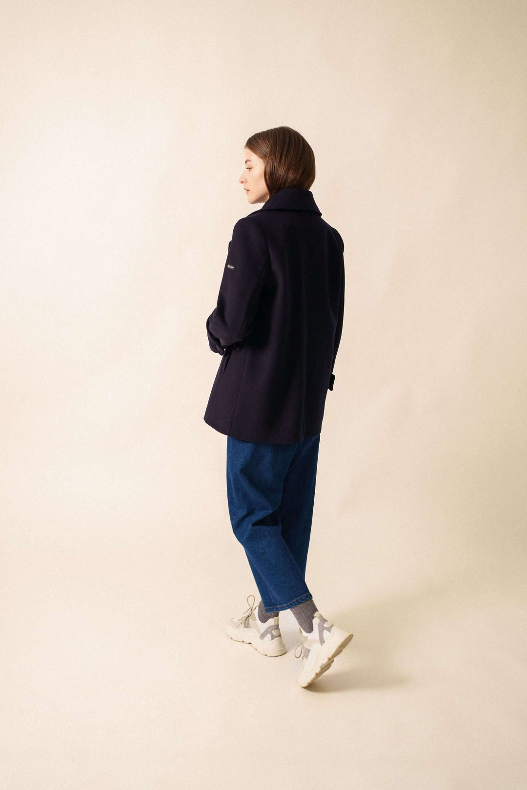 Femme portant le manteau chic STE ROMY, longueur sous les fesses avec col en V, deux poches et boutons, sur un fond neutre.