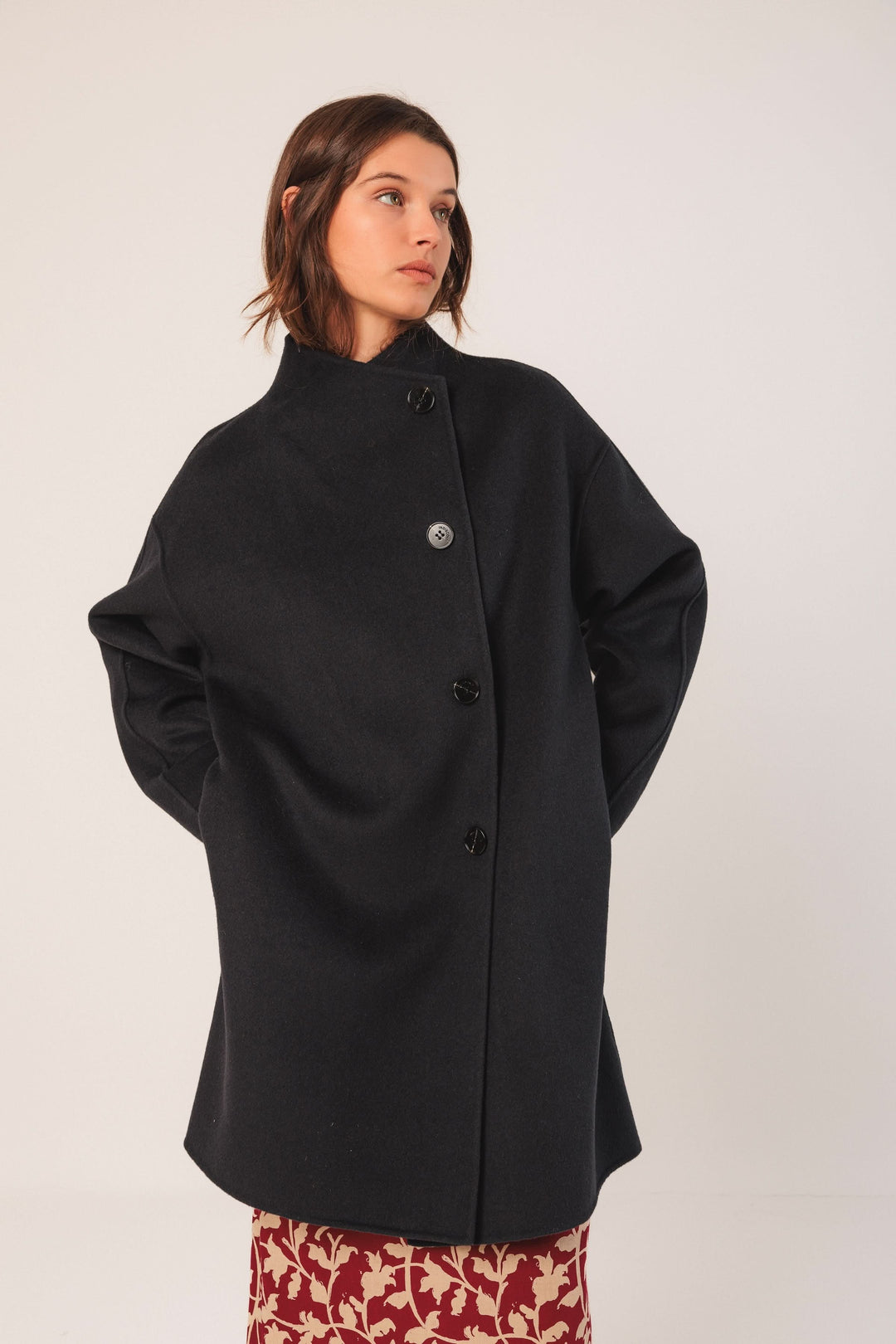 Manteau minimaliste noir avec coupe ample, col montant et fermeture boutonnée, porté sur une jupe imprimée.